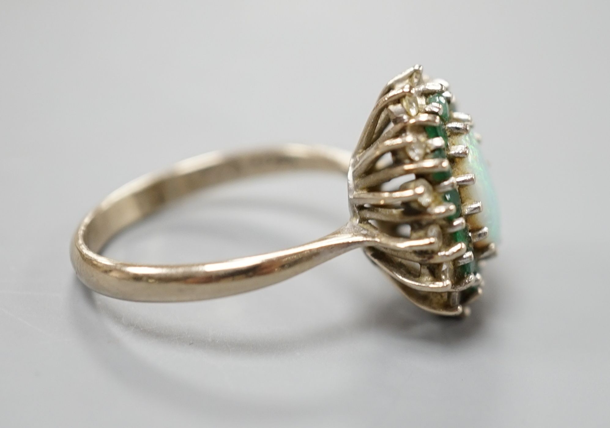 An 18ct white metal, white opal, emerald and diamond set oval cluster ring, size O, gross weight 6.2 grams.